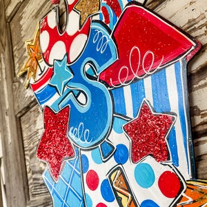 May include: A wooden door hanger decorated with red, white and blue stars, stripes and polka dots. The hanger is shaped like the letters "USA" and features a rocket ship design with a sparkly finish.