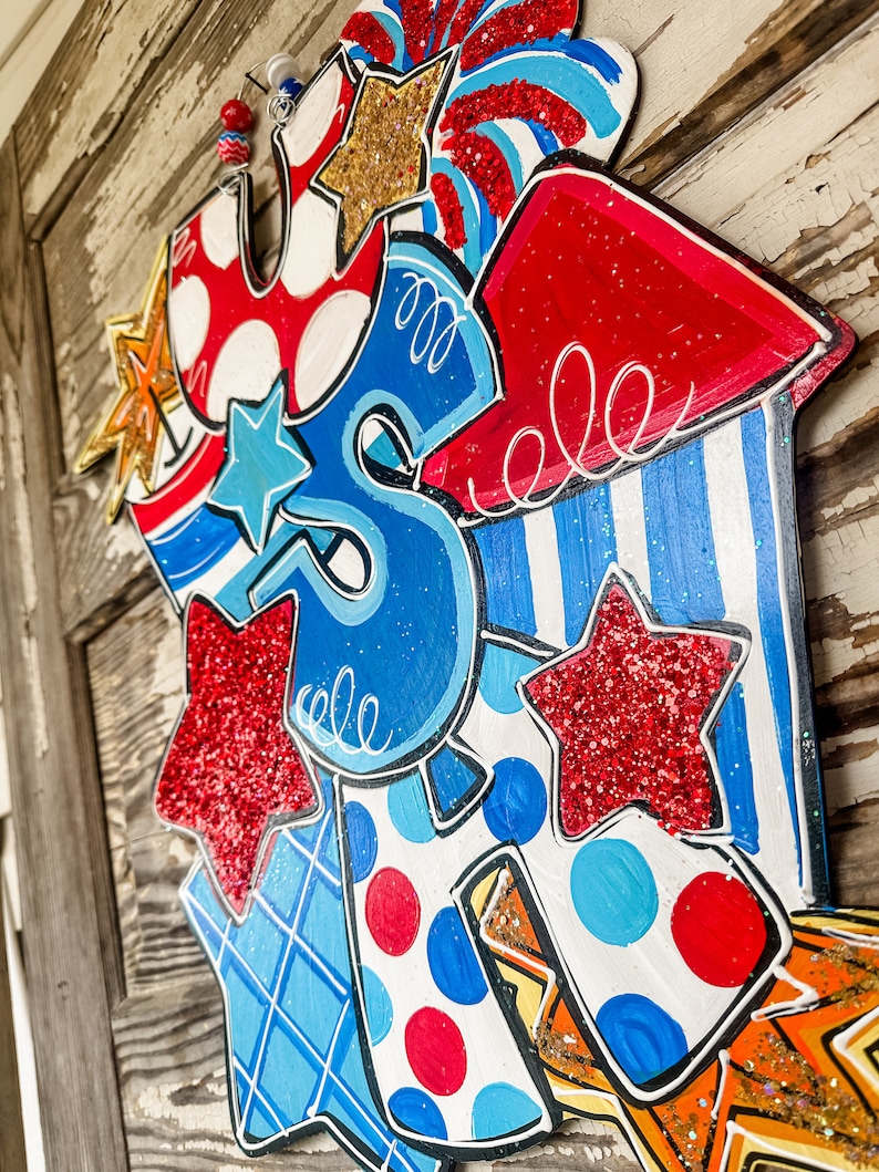 May include: A wooden door hanger decorated with red, white and blue stars, stripes and polka dots. The hanger is shaped like the letters "USA" and features a rocket ship design with a sparkly finish.