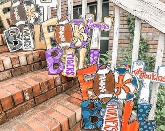 May include: A collection of wooden football-themed signs with different team names and colors. The signs are decorated with pom-poms, footballs, and stripes. The signs are arranged in a staircase pattern.