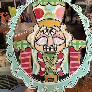 Puede incluir: Decoración de cascanueces de madera pintada a mano. El diseño presenta una figura de cascanueces sonriente con un sombrero dorado, una chaqueta a rayas rojas y verdes y un borde festoneado azul claro. El fondo está borroso.