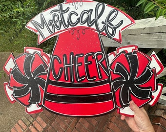 May include: Red and black wooden cheerleading megaphone door hanger with the text "Metcalf Cheer" and two black and white pom-poms.