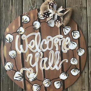 May include: Round wooden door hanger with a brown background and white cotton boll garland. The words "Welcome Y'all" are painted in gold and white on the door hanger. A brown and white bow is attached to the top of the door hanger.