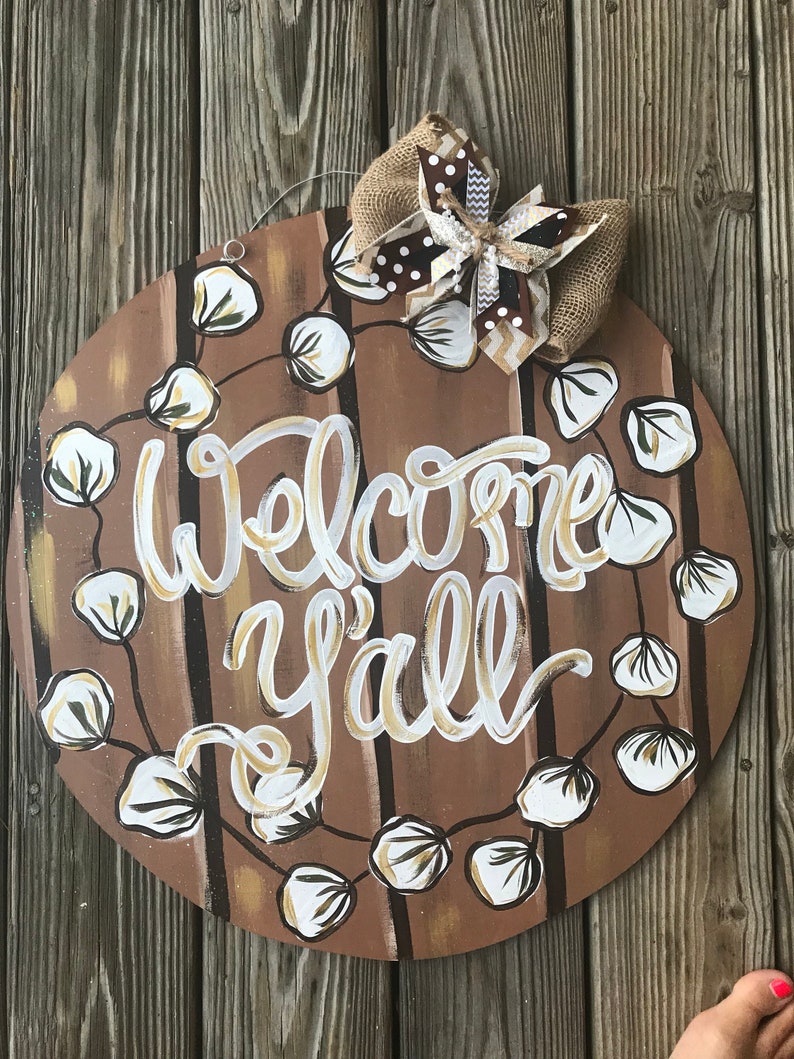 May include: Round wooden door hanger with a brown background and white cotton boll garland. The words "Welcome Y'all" are painted in gold and white on the door hanger. A brown and white bow is attached to the top of the door hanger.