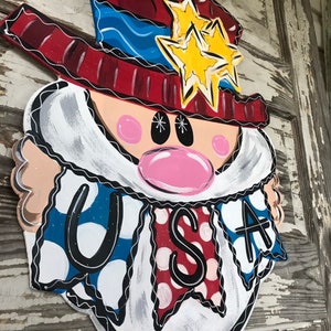 May include: A wooden door hanger shaped like a cartoon face with a red, white, and blue hat, a white beard, and a banner that says "USA".