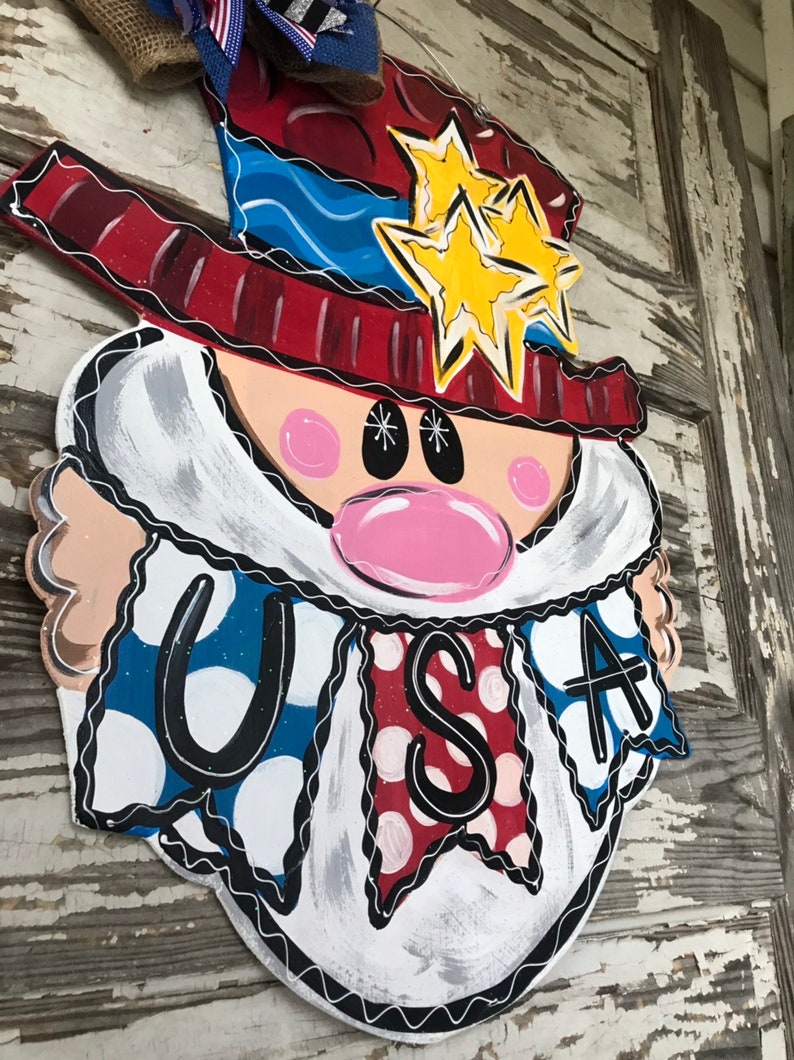 May include: A wooden door hanger shaped like a cartoon face with a red, white, and blue hat, a white beard, and a banner that says "USA".