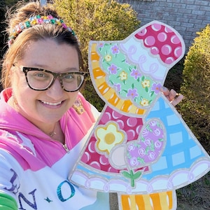 May include: A colorful, wooden Easter decoration in the shape of a bonnet. The bonnet features a patchwork design with pink polka dots, floral patterns, and a blue checkered section. The edges are outlined with a white stitch effect.