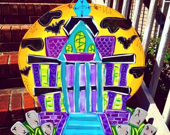 May include: A hand-painted wooden Halloween sign featuring a purple and green haunted house with a blue door and yellow moon. The sign is decorated with bats and a compass.