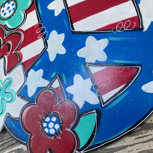 May include: A blue and white peace sign with red, white, and blue stars and stripes. The peace sign is decorated with flowers in red, white, and green.