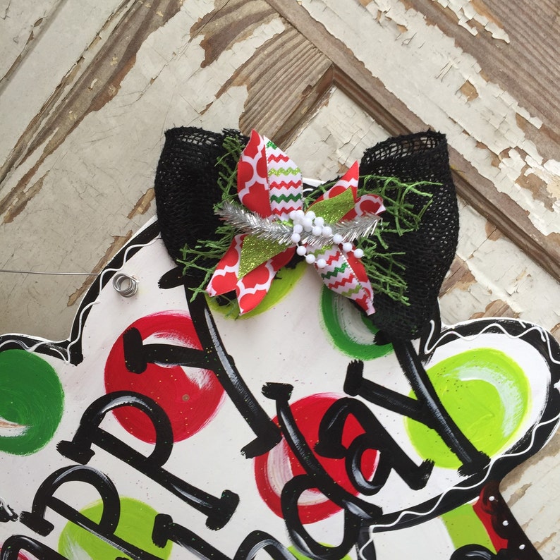 May include: A white wooden door hanger shaped like a mitten with red and green polka dots. The mitten has the words "Happy Holidays" written on it in black. A black and green bow with a silver and white ribbon is attached to the top of the mitten.