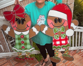 May include: A woman holds up two large gingerbread people cutouts. The gingerbread person on the left is a girl with a red bow and a green and white plaid dress. The gingerbread person on the right is a boy with a red and white Santa hat and a red and white plaid shirt.
