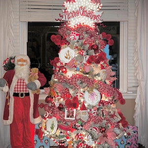 May include: A festive Christmas tree adorned with red and white decorations, fairy lights, and a "Merry Christmas" sign. A Santa Claus figure stands beside it, holding a teddy bear. Presents are at the base.