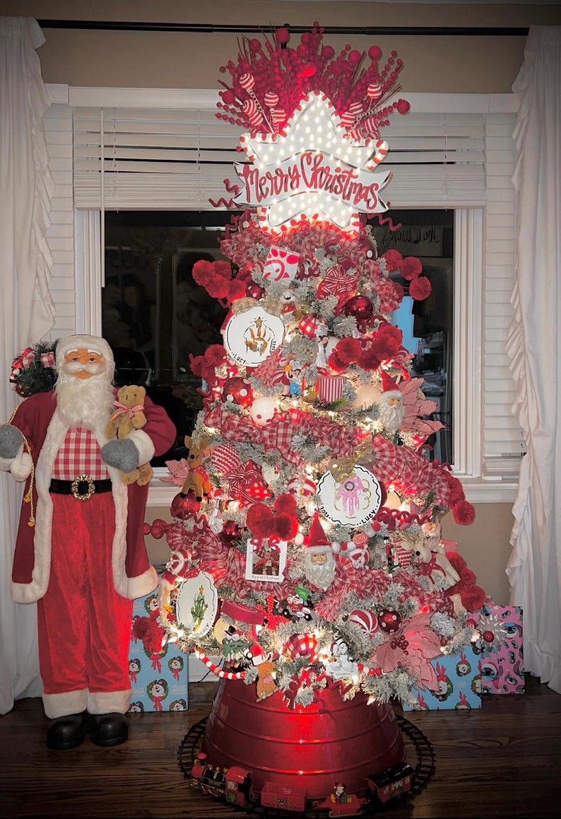May include: A festive Christmas tree adorned with red and white decorations, fairy lights, and a "Merry Christmas" sign. A Santa Claus figure stands beside it, holding a teddy bear. Presents are at the base.