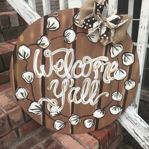 May include: A round wooden door hanger with a brown and white striped background. The words "Welcome Y'all" are written in white paint. The door hanger is decorated with a brown burlap bow and white cotton boll accents.