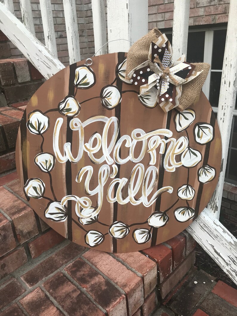 May include: A round wooden door hanger with a brown and white striped background. The words "Welcome Y'all" are written in white paint. The door hanger is decorated with a brown burlap bow and white cotton boll accents.