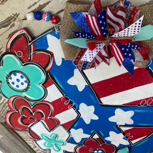 May include: A patriotic peace sign door hanger with red, white, and blue flowers and a star-spangled bow. The peace sign is painted in blue with white stars and red and white stripes.