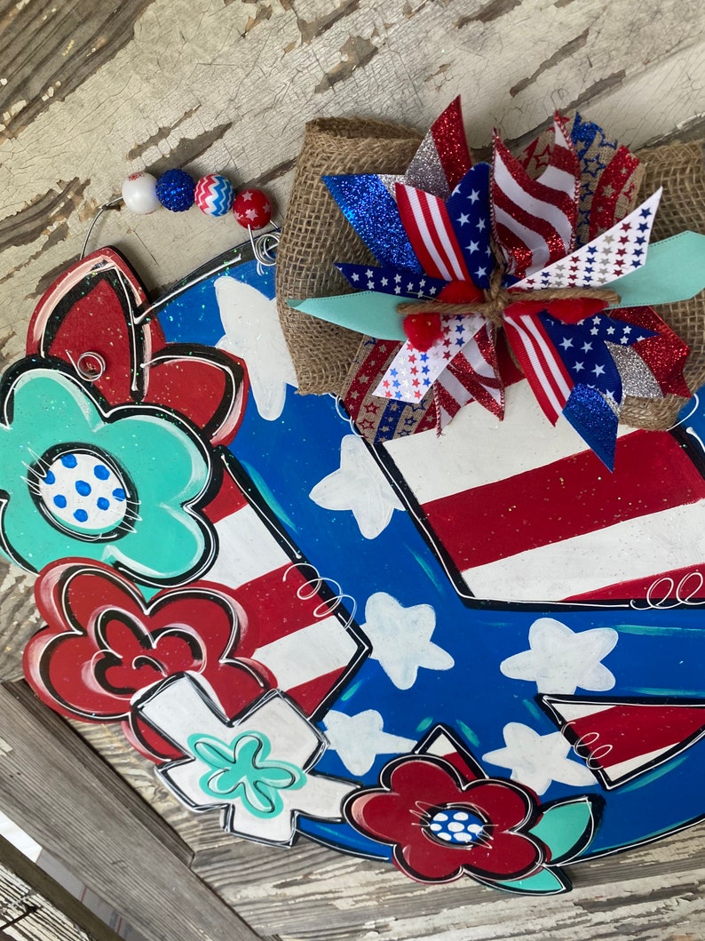 May include: A patriotic peace sign door hanger with red, white, and blue flowers and a star-spangled bow. The peace sign is painted in blue with white stars and red and white stripes.