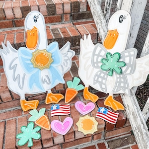 May include: Two wooden duck decorations with white bodies, orange beaks and feet, and large bows. One bow is light blue, the other is gray. Additional wooden cutouts include carrots, hearts, shamrocks, and American flags. The decorations are on a brick surface.