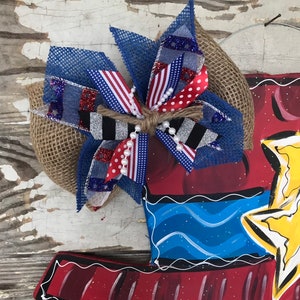 May include: A red, white, and blue bow with burlap and glitter accents. The bow is tied with twine and has white pearl beads. The bow is attached to a wooden door hanger with a painted design of a person wearing a hat and a star.