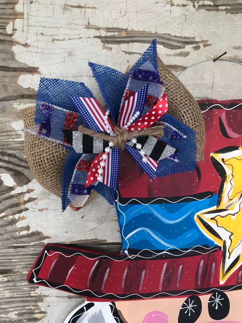 May include: A red, white, and blue bow with burlap and glitter accents. The bow is tied with twine and has white pearl beads. The bow is attached to a wooden door hanger with a painted design of a person wearing a hat and a star.