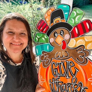 Könnte beinhalten: Ein bunter hölzerner Truthahn-Türhänger mit einer Kürbisform als Basis. Der Truthahn trägt einen Pilgerhut und hat einen roten Schnabel. Der Kürbis hat die Worte "Give thanks" darauf geschrieben.