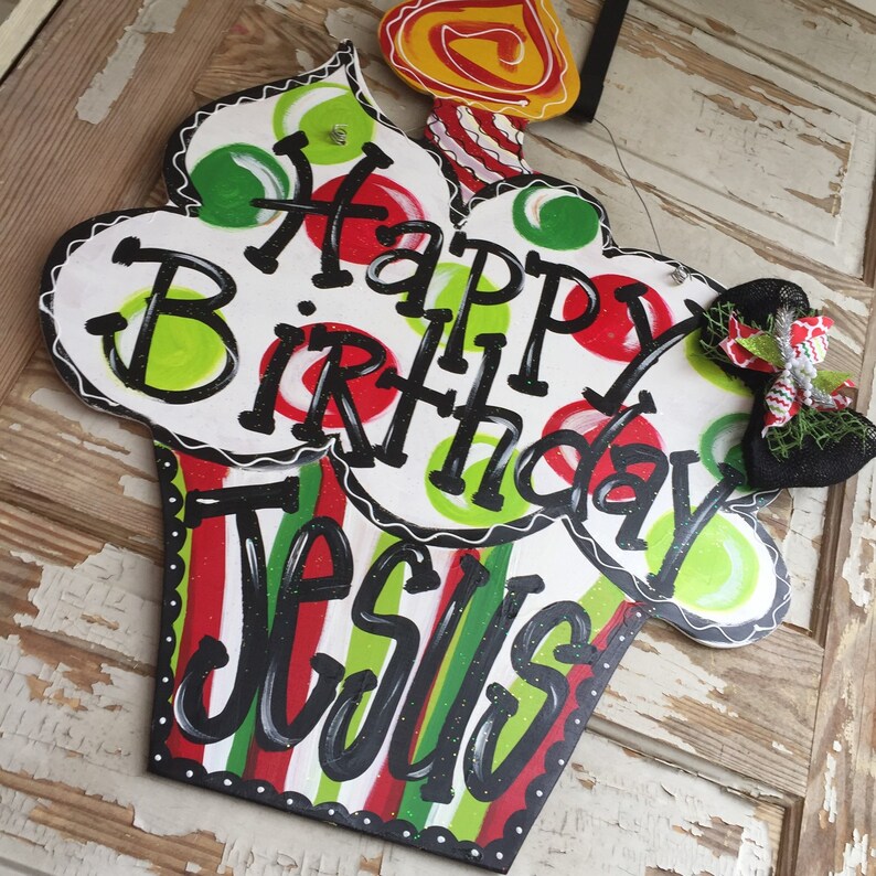 May include: A white cupcake-shaped wooden sign with red, green, and white stripes. The sign says "Happy Birthday Jesus" in black lettering with green and red polka dots. A yellow candle with red and white stripes is on top of the cupcake.