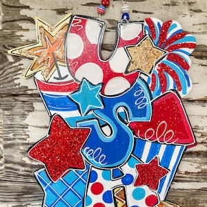 May include: A wooden door hanger featuring the letters "USA" in red, white and blue with glitter stars and other patriotic designs.
