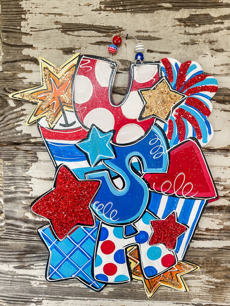 May include: A wooden door hanger featuring the letters "USA" in red, white and blue with glitter stars and other patriotic designs.