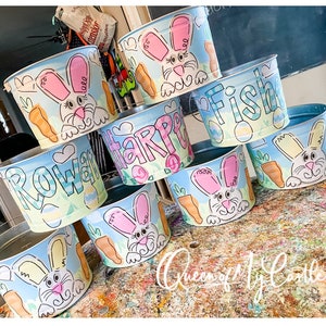May include: A collection of painted metal buckets with Easter-themed designs. Each bucket features a cartoon bunny, carrots, and pastel colors. Some buckets have names like "Rowan", "Harper", and "Fish" painted on them. The buckets are arranged on a colorful surface.