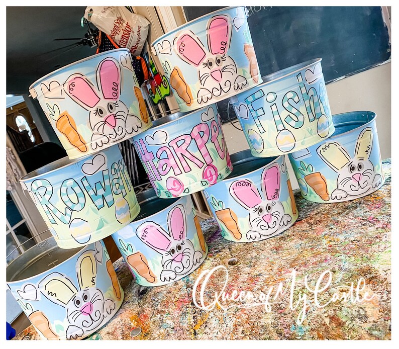 May include: A collection of painted metal buckets with Easter-themed designs. Each bucket features a cartoon bunny, carrots, and pastel colors. Some buckets have names like "Rowan", "Harper", and "Fish" painted on them. The buckets are arranged on a colorful surface.