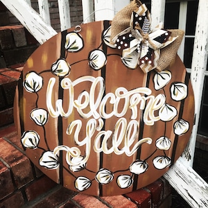 May include: A round wooden door hanger with a brown and white striped background. The words "Welcome Y'all" are painted in white with a gold outline. A brown and white bow with a white cotton ball garland surrounds the text.