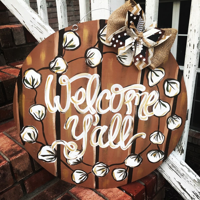 May include: A round wooden door hanger with a brown and white striped background. The words "Welcome Y'all" are painted in white with a gold outline. A brown and white bow with a white cotton ball garland surrounds the text.