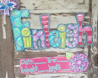May include: A colorful wooden name sign with the name "Finleigh" painted in bright colors with polka dots and stripes. A pink and green sign below has the words "Date: Time: Weight: Length:" with blank lines for filling in baby stats.