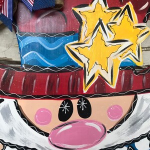 May include: A wooden door hanger featuring a cartoon Uncle Sam with a red, white, and blue hat, a blue and white striped shirt, and a red, white, and blue bow tie. The hanger has two yellow stars on the hat and the words "USA" at the bottom.
