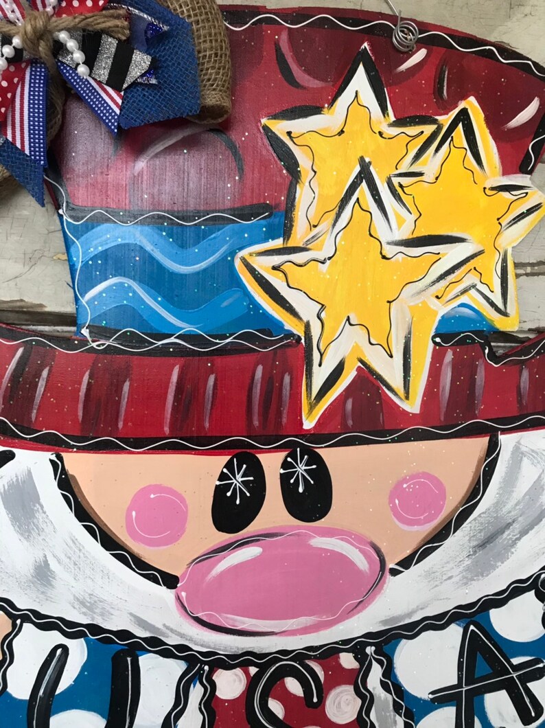 May include: A wooden door hanger featuring a cartoon Uncle Sam with a red, white, and blue hat, a blue and white striped shirt, and a red, white, and blue bow tie. The hanger has two yellow stars on the hat and the words "USA" at the bottom.