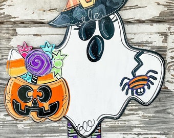 May include: A white ghost wearing a black witch hat with a purple and orange striped bow, holding a jack-o-lantern with candy corn and a purple flower. The ghost has black eyes and a spider on its side. The ghost has striped legs and black boots.