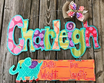 May include: A wooden name sign with the name "Charleigh" in colorful letters with polka dots and stripes. Below the name is a wooden sign with an elephant illustration and the words "Date: Time: Weight: Length:"