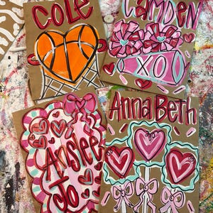 Op de afbeelding: Vier bruine papieren cadeauzakken met handgeschilderde ontwerpen. E&eacute;n zak heeft een basketbal in een hart, een andere heeft bloemen en "XO", en de andere hebben namen en harten. De zakken zijn versierd met rode, roze en blauwe verf.
