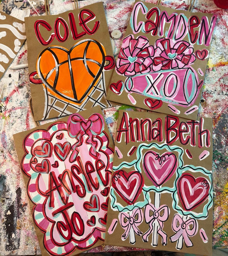 Op de afbeelding: Vier bruine papieren cadeauzakken met handgeschilderde ontwerpen. E&eacute;n zak heeft een basketbal in een hart, een andere heeft bloemen en "XO", en de andere hebben namen en harten. De zakken zijn versierd met rode, roze en blauwe verf.