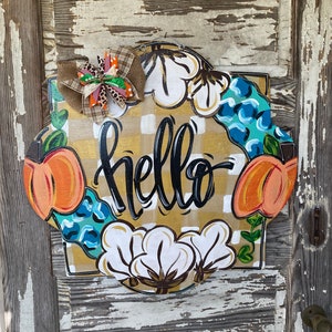 May include: A round, wooden door hanger with a yellow and white checkered background. The word "hello" is written in black script. The wreath is decorated with cotton bolls, peach-colored pumpkins, and blue and green leaves.