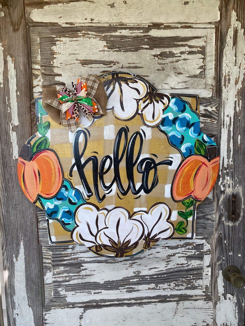 May include: A round, wooden door hanger with a yellow and white checkered background. The word "hello" is written in black script. The wreath is decorated with cotton bolls, peach-colored pumpkins, and blue and green leaves.