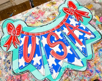 May include: A wooden sign shaped like a banner with the word "USA" in red letters on a blue and white star pattern. The banner is decorated with red bows and a turquoise background.
