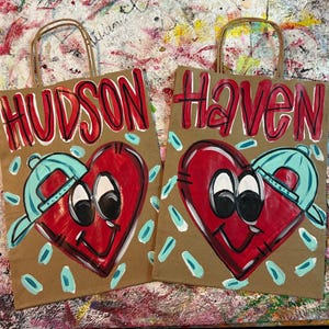 Op de afbeelding: Twee bruine papieren cadeauzakken met rode letters met de namen "HUDSON" en "Haven". Elke tas heeft een rood hart met een blauwe baseballpet. De harten hebben zwarte ogen en een lachende mond. Blauwe en witte accenten omringen de harten.