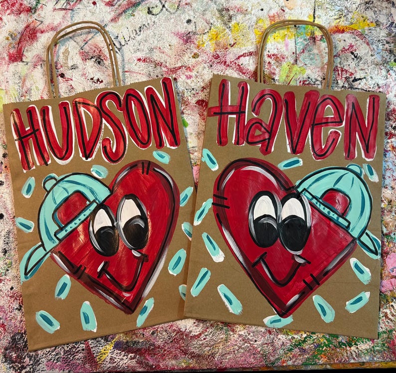 Op de afbeelding: Twee bruine papieren cadeauzakken met rode letters met de namen "HUDSON" en "Haven". Elke tas heeft een rood hart met een blauwe baseballpet. De harten hebben zwarte ogen en een lachende mond. Blauwe en witte accenten omringen de harten.