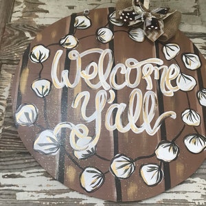 May include: A round wooden door hanger with a brown background and white and gold lettering that says "Welcome Y'all". The door hanger is decorated with a garland of white cotton balls and a brown and white bow.