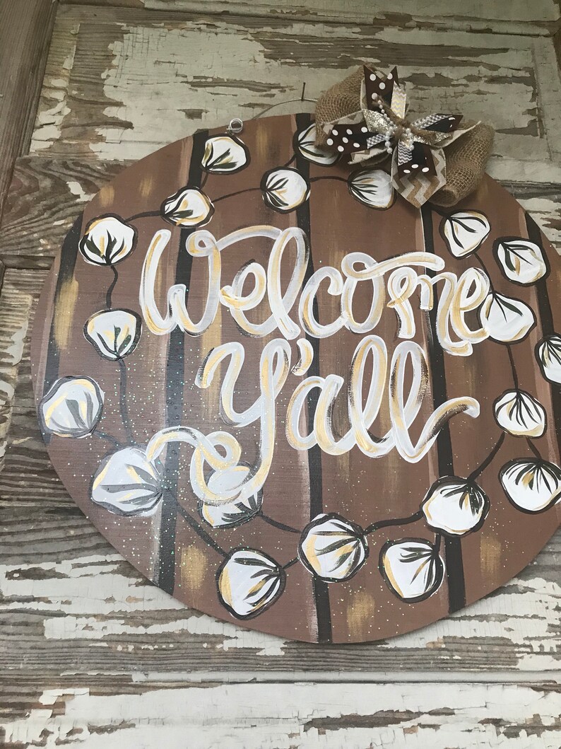 May include: A round wooden door hanger with a brown background and white and gold lettering that says "Welcome Y'all". The door hanger is decorated with a garland of white cotton balls and a brown and white bow.