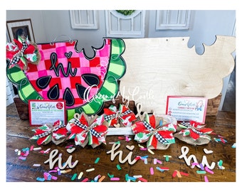 May include: A watermelon-shaped wooden door hanger with the word "hi" painted on it. The hanger is decorated with pink and green checkered patterns. There are four smaller watermelon-themed bows on a wooden table with confetti and the word "hi" written in wood.