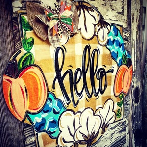 May include: A wooden door hanger with a checkered background and the word "hello" in black script. The hanger is decorated with painted pumpkins, cotton bolls, and blue and green leaves. A brown and leopard print bow is attached to the top.