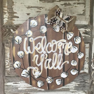 May include: A round wooden door hanger with a brown background and white cotton boll garland. The words "Welcome Y'all" are painted in gold on the door hanger. A brown and white burlap bow is attached to the top of the door hanger.