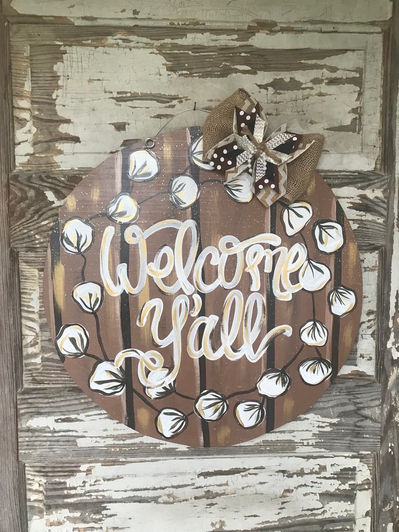 May include: A round wooden door hanger with a brown background and white cotton boll garland. The words "Welcome Y'all" are painted in gold on the door hanger. A brown and white burlap bow is attached to the top of the door hanger.