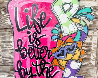 Pode incluir: Uma placa de madeira rosa e branca com o texto "Life is better by the" e uma ilustração colorida de uma cena de praia com um sol, um coração e as letras "P" e "L".